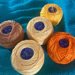 May include: Five spools of Cebelia DMC thread in various colors: light brown, yellow, and orange. Each spool has a blue label with the brand name and thread weight. The threads are arranged on a blue surface.