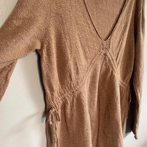 Puede incluir: Vestido marr&oacute;n claro de manga larga con cuello en V. El vestido presenta detalles de cord&oacute;n en la cintura y las mangas, y un elemento decorativo en el escote. La tela tiene una apariencia texturizada.