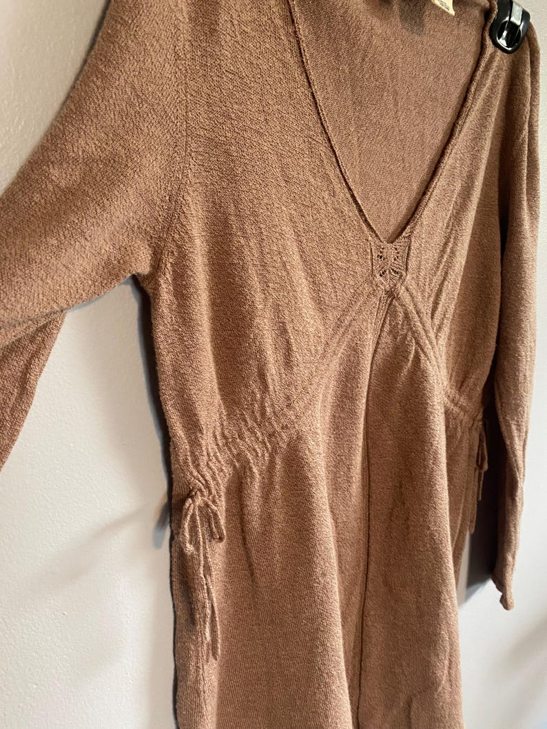 Puede incluir: Vestido marr&oacute;n claro de manga larga con cuello en V. El vestido presenta detalles de cord&oacute;n en la cintura y las mangas, y un elemento decorativo en el escote. La tela tiene una apariencia texturizada.