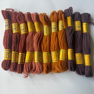 May include: A collection of DMC embroidery floss in various shades of brown, rust, and gold. Each skein is labeled with a yellow band displaying the DMC logo and color number. The floss is arranged neatly, showcasing the range of colors available for needlework projects.