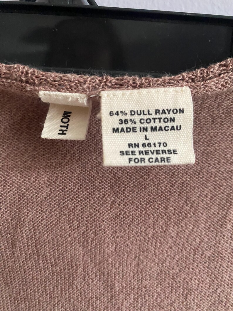 Puede incluir: Primer plano de una etiqueta de prenda de color malva con la marca "MOTH". La etiqueta incluye la composici&oacute;n de la tela: 64% ray&oacute;n opaco, 36% algod&oacute;n, y el texto "MADE IN MACAU". La talla es L, con RN 66170 e instrucciones de cuidado.