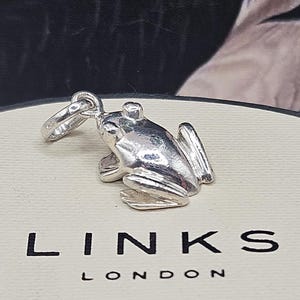May include: A polished silver frog charm with a small circular jump ring attached to its head. The charm is positioned on a cream-coloured circular surface featuring the black text LINKS LONDON printed in a clean, sans-serif font.