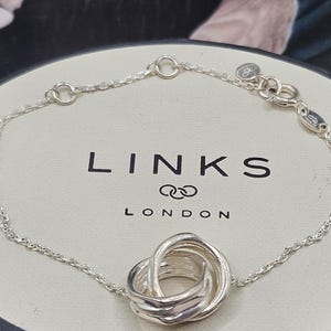May include: A silver bracelet featuring a woven chain and a central interlocking knot design. The bracelet is presented on a white box with the brand name "LINKS LONDON" printed in black.