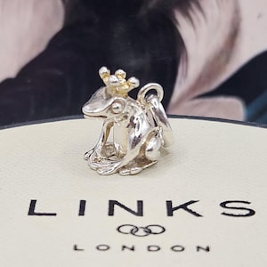 Könnte beinhalten: Ein silberner Frosch-Anhänger mit goldener Krone. Der Frosch ist detailliert mit sichtbaren Augen und Beinen. Der Anhänger liegt auf einer weißen Oberfläche mit dem schwarzen Aufdruck "LINKS LONDON".