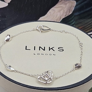 Può includere: Un braccialetto d'argento con un ciondolo a forma di cuore e ciondoli geometrici. Il braccialetto è su una scatola bianca con il testo "LINKS LONDON" su di essa.