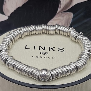 May include: A silver-toned stretch bracelet composed of numerous small, rounded metal rings strung together. One central bead features the engraved text LINKS LONDON. The bracelet is displayed on a circular cream-coloured stand with black trim.