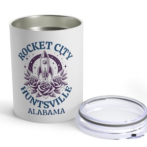 May include: White stainless steel travel mug with a blue and purple design. The design features a rocket ship, roses, and the text "Rocket City Huntsville Alabama".