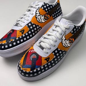 May include: A pair of white sneakers with black and white checkered patterns, yellow and orange accents, and hand-painted designs. The designs include a red and blue building, a yellow and orange cloud, and a brown cigar.