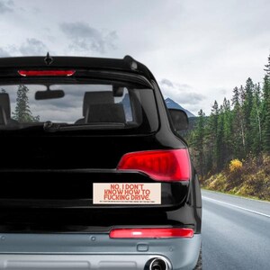 May include: Black SUV with a bumper sticker that reads "NO, I DON'T KNOW HOW TO FUCKING DRIVE." The sticker is rectangular with red text on a beige background. The car is on a road with trees and mountains in the background.