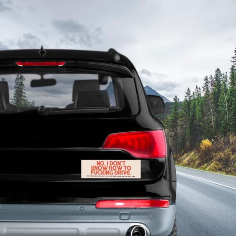 May include: Black SUV with a bumper sticker that reads "NO, I DON'T KNOW HOW TO FUCKING DRIVE." The sticker is rectangular with red text on a beige background. The car is on a road with trees and mountains in the background.
