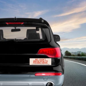 May include: A black SUV with a bumper sticker that reads "NO, I DON'T KNOW HOW TO FUCKING DRIVE." The sticker has red text on a beige background. The car is on a road with a blue sky.