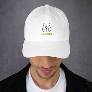 May include: White baseball cap with a curved peak. The cap has a navy blue bear graphic and the word "CALIFORNIA" embroidered in gold thread. The cap is worn by a person.