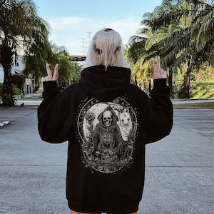 Puede incluir: Una sudadera con capucha negra con un estampado gráfico de un esqueleto sentado en un trono con una luna creciente detrás. La sudadera tiene capucha y cremallera.
