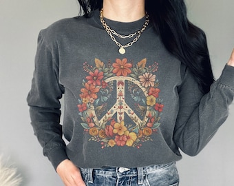 Può includere: Maglietta a maniche lunghe grigio scuro con un design a simbolo della pace circondato da fiori colorati. Il simbolo della pace è al centro della ghirlanda floreale. La maglietta è abbinata a jeans blu.