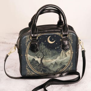 May include: Black handbag with a celestial design featuring a tree, mountains, moon, and stars. The bag has a black handle and a detachable shoulder strap with gold-coloured hardware. The bag is approximately 23 cm wide.