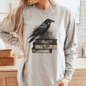 May include: A gray long-sleeve t-shirt with a black crow perched on a stack of books. The crow is facing the right side of the image. The shirt has a distressed, vintage look.