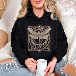 Può includere: Felpa con cappuccio nera con un design dettagliato di libellula nei toni del marrone e dell'oro. La libellula è centrata all'interno di una cornice circolare ornata con accenti floreali. La felpa ha un cappuccio con coulisse e una tasca a marsupio.