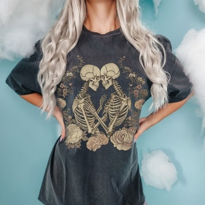 Op de afbeelding: Een zwart oversized T-shirt met een grafische print van twee skeletten die elkaar omhelzen, omringd door bloemdetails. De skeletten zijn lichtbruin van kleur.