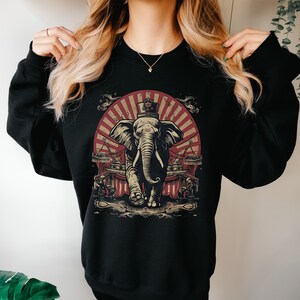 May include: Black sweatshirt with a graphic print of an elephant wearing a top hat and standing in front of a red and white striped tent. The elephant is surrounded by other circus performers.