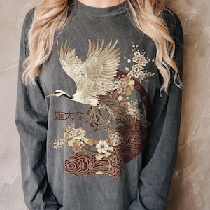 Puede incluir: Camiseta de manga larga gris oscuro con una ilustración detallada de una grulla en vuelo, rodeada de motivos florales y de olas. El diseño incluye texto japonés. La grulla tiene plumas doradas y blancas, sobre un fondo burdeos y marrón.