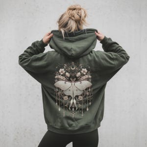 Puede incluir: Una sudadera con capucha verde oliva con un diseño de polilla y flores en la espalda. La polilla es de color gris claro con detalles, rodeada de flores y una luna creciente. La sudadera tiene una capucha con cordón y un ajuste relajado.
