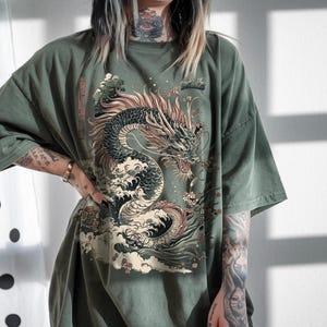 May include: An oversized green t-shirt featuring a graphic print of a black dragon in a wave with pink flowers. The dragon is facing the right side of the image.