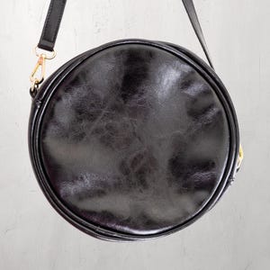 May include: A black, round crossbody bag with a leather-like texture. The bag has a long, adjustable strap and gold-colored hardware. The bag is circular in shape and appears to be made of a durable material.