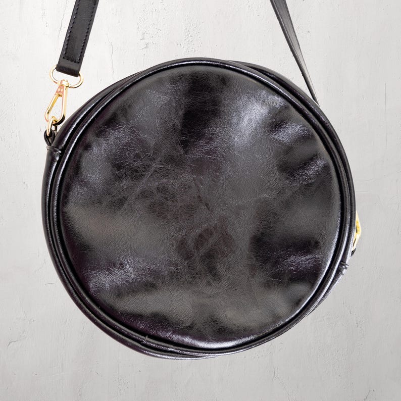 May include: A black, round crossbody bag with a leather-like texture. The bag has a long, adjustable strap and gold-colored hardware. The bag is circular in shape and appears to be made of a durable material.