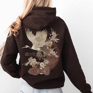 Könnte beinhalten: Ein braunes Hoodie mit einem grafischen Aufdruck einer fliegenden Kranich mit Blumen und dem japanischen Text "雄大なる" auf der Rückseite.