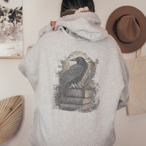 May include: A gray hooded sweatshirt with a black raven perched on a stack of books. The raven is surrounded by a distressed, faded design. The text "NEVERMORE" is printed vertically on the left side of the design.