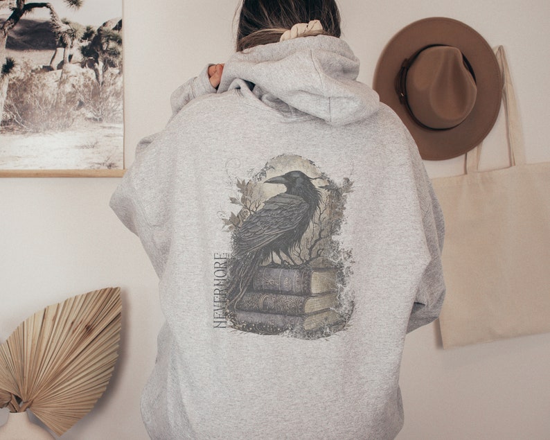 May include: A gray hooded sweatshirt with a black raven perched on a stack of books. The raven is surrounded by a distressed, faded design. The text "NEVERMORE" is printed vertically on the left side of the design.