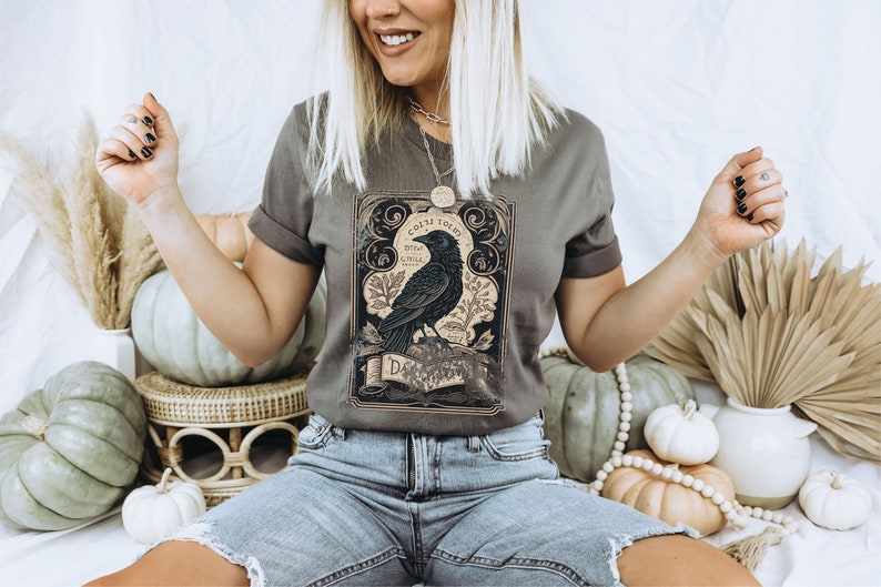 Peut inclure: Un t-shirt gris avec une illustration vintage d'un corbeau et le texte "Once Told" dans une police d&eacute;corative. Le corbeau est perch&eacute; sur une branche avec des feuilles et des fleurs. Le t-shirt est port&eacute; par une personne assise sur une surface blanche avec des citrouilles et d'autres d&eacute;corations d'automne.