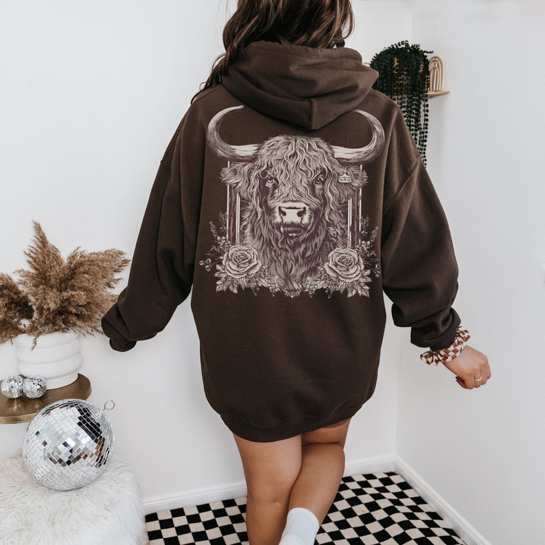 May include: A dark brown hoodie featuring a detailed illustration of a Highland cow on the back. The cow is framed with vertical lines and surrounded by roses. The hoodie has a hood and long sleeves.