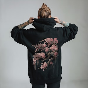May include: Black hoodie with a cherry blossom tree design on the back. The illustration features pink flowers and brown branches. The hoodie has a drawstring hood and long sleeves.