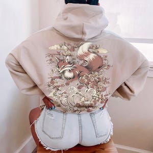 May include: A beige hoodie featuring a graphic print of a fox surrounded by flowers and waves. The fox is brown and white with a bushy tail.