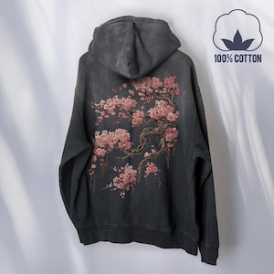 May include: Dark grey hoodie with a floral design on the back. The design features pink cherry blossoms and brown branches. The hoodie is made of 100% cotton.