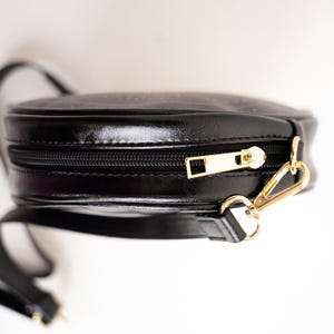 May include: Black, round crossbody bag with a gold zipper and clasp. The bag has a long, black strap and a shiny, leather-like finish. The zipper pull and clasp are gold-colored.