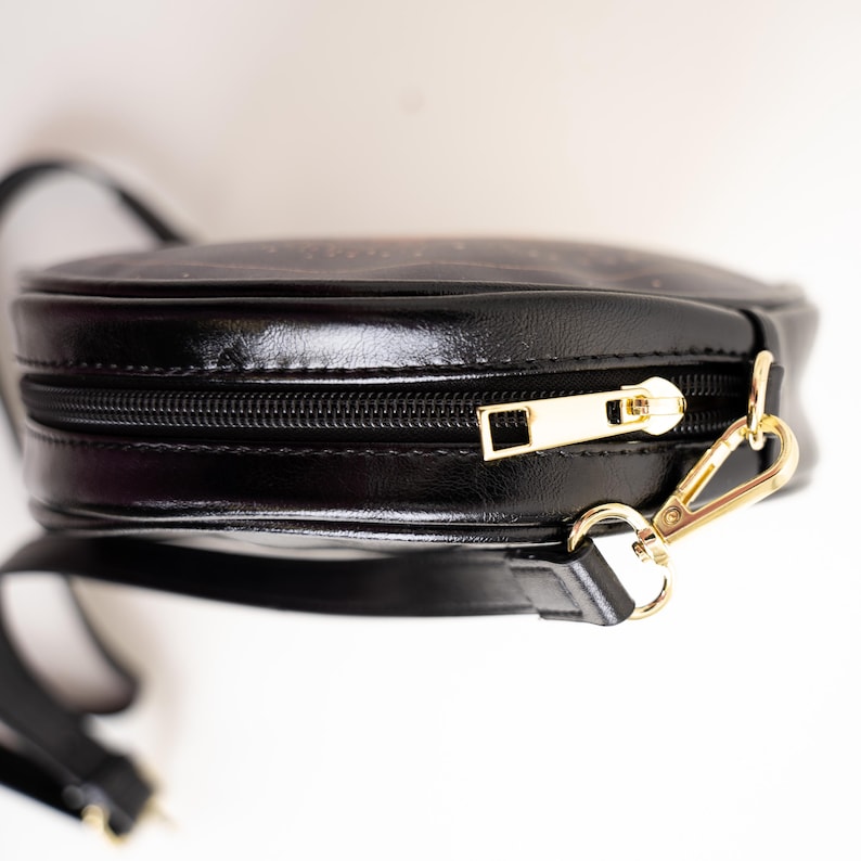 May include: Black, round crossbody bag with a gold zipper and clasp. The bag has a long, black strap and a shiny, leather-like finish. The zipper pull and clasp are gold-colored.
