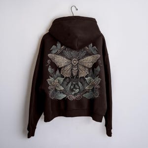 May include: A dark brown hoodie featuring a detailed moth and floral design on the back. The moth has intricate wing patterns, surrounded by leaves and flowers in shades of green and grey. The hoodie is suspended against a white backdrop.
