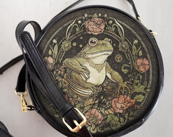 May include: A black round crossbody bag with a long adjustable strap. The bag features a circular design with a detailed illustration of a frog sitting in a floral arrangement on a black background.