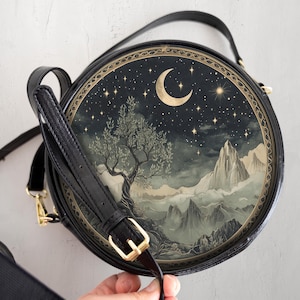 May include: A black round crossbody bag with a gold trim and a crescent moon and stars design. The bag features a landscape scene with a tree and mountains.