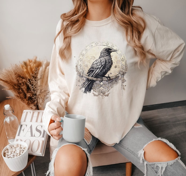 Pu&ograve; includere: Una felpa a maniche lunghe color crema con un'illustrazione di corvo nero in una cornice circolare. Il corvo &egrave; appollaiato su un ramo con intricati dettagli floreali. La felpa ha una vestibilit&agrave; rilassata ed &egrave; perfetta per l'abbigliamento casual.