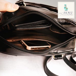 K&ouml;nnte beinhalten: Schwarze Leder-Umh&auml;ngetasche mit oberem Griff, ge&ouml;ffnet, um ein Portemonnaie und ein goldfarbenes Smartphone zu zeigen. Die Tasche hat einen Rei&szlig;verschluss und einen langen Riemen. Das Logo "Jenu Trinkets" befindet sich oben rechts.