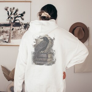 May include: White hooded sweatshirt with a black raven graphic on the back. The graphic features a raven perched on a stack of books with the word "Nevermore" printed vertically on the left side of the graphic.