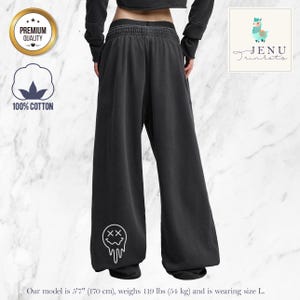 Può includere: Pantaloni della tuta neri a gamba larga con una grafica bianca di una faccia che si scioglie sulla gamba sinistra. I pantaloni sono realizzati in 100% cotone e hanno una vestibilità ampia. L'immagine include anche un distintivo "Premium Quality".