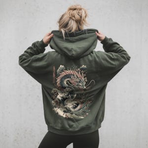 Könnte beinhalten: Ein grüner Kapuzenpullover mit einem grafischen Aufdruck eines schwarzen Drachen, der aus Wellen aufsteigt. Der Drache ist von rosa Blumen und einem japanischen Schriftzeichen umgeben.
