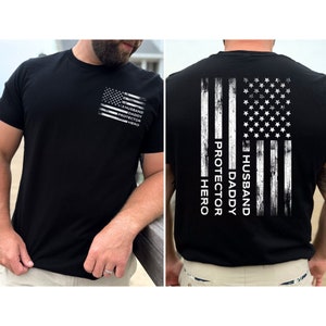 May include: Black t-shirt with a distressed American flag design on the back. The flag has the words "Husband, Daddy, Protector, Hero" written vertically down the left side. The front of the shirt has a smaller distressed American flag with the same words written vertically on the right side.