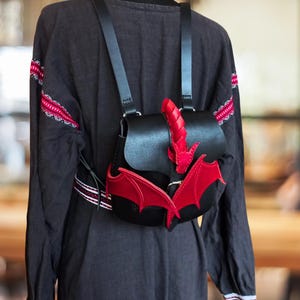 Pode incluir: Uma mochila de couro preto com asas e cauda de dragão vermelhas. A mochila tem alças ajustáveis e um fecho de aba.