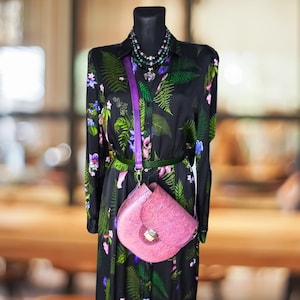 May include: A black floral dress with a purple strap and a pink leather satchel bag. The dress has a green belt and a layered necklace with a heart pendant.