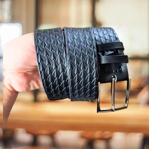 May include: A black belt featuring a repeating embossed geometric pattern of raised triangular spikes across the strap. The belt includes a smooth black leather keeper loop and a polished silver-toned metal rectangular pin buckle.